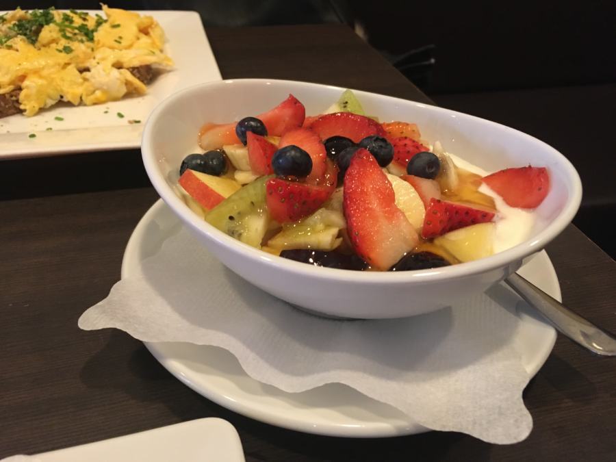 Ovale Schüssel mit Joghurt und frischen Erdbeeren, Heidelbeeren, Kiwi und Äpfeln. Im Hintergrund sieht man einen eckigen weißen Teller mit Eierspeisbrot