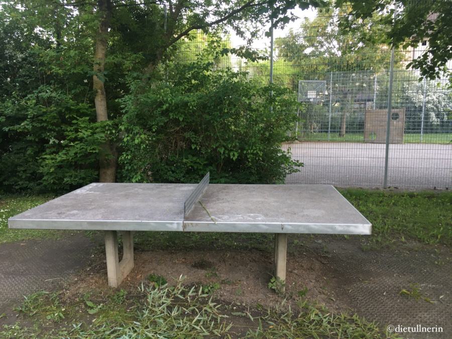 Tischtennisplatte am Spielplatz. im Hintergrund ist ein eingezäunter Ballspiele-Platz zu sehen 