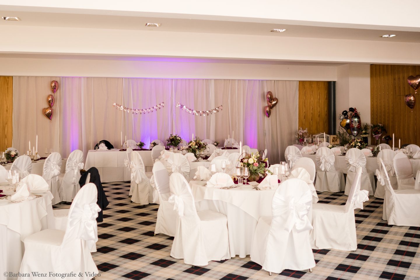Der Festsaal des Berghotels Tulbinger Kogel. Festlich dekoriert für eine Hochzeit. Tische weiß gedeckt, Stühle mit weißen Stuhlhussen mit Satinmaschen an der Lehne. Der Teppichboden ist weiß-grau-schwarz kariert. Im Hintergrund rosa Luftballons.