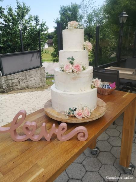 4-stöckige weiße Hochzeitstorte mit rosa Rosen, Eukalyptusblättern und weißem Schleierkraut dekoriert. Die Torte steht auf einem braunen Holztisch, im Vordergrund ist eine rosa Holzdeko/Schriftzug "love" zu sehen. Im Hintergrund sieht man eine Garteneinfahrt, in der der Tisch steht.
