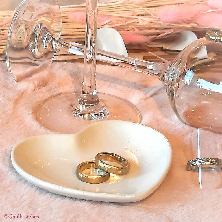 hellrosa Stoff aus Fellimitat als Tischtuch mit einem stehenden und einem liegenden Weinglas im Hintergrund. Im Vordergrund liegen auf einem herzförmigen Teller 2 Eheringe in Gelbgold. Rechts daneben sieht man einen weiteren Ring in Weißgold.