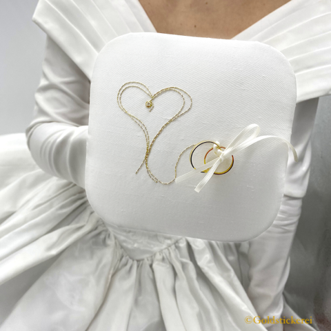 Zu sehen ist eine Braut im weißen Brautkleid, die ein weißes Ringkissen zeigt. Das Kissen ist mit Goldfäden bestickt. Und zwar sieht man zwei Herzen und ein weißes Satinband, mit dem die beiden Eheringe am Kissen befestigt sind.