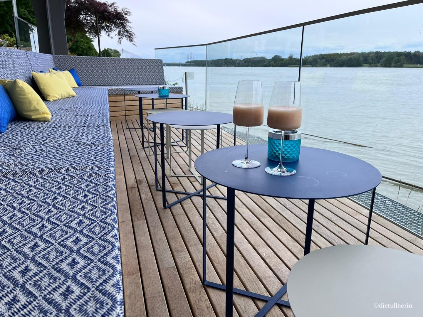 Terrasse des Restaurants Süddeck in Tulln mit blau-weiß-gemusterten Sitzauflagen, runden Metalltischen und edlem Boden in Holzstyle. Direkt am Ufer der Donau.