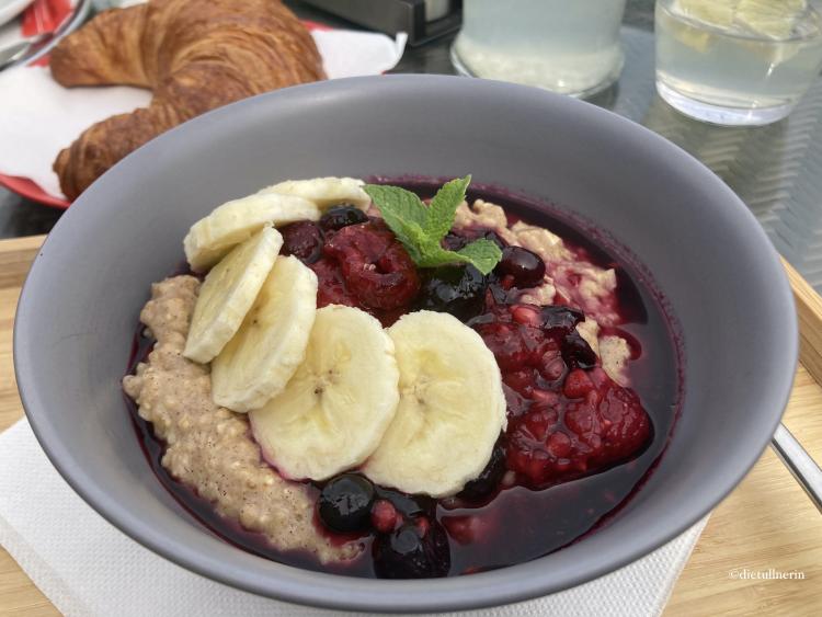 Porridge mit Banane und Waldbeeren in grauer Schüssel, Croissant im Hintergrund zu sehen