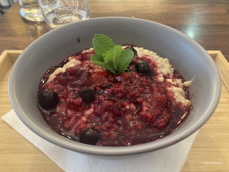 ayurvedischer Porridge mit Himbeeren und Heidelbeeren sowie Minzblättchen zur Deko. Angerichtet in grauer Schüssel auf Holztablett.